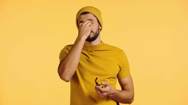 Joven cansado en gorro sombrero frotando los ojos y sosteniendo gafas aisladas en amarillo - foto de stock