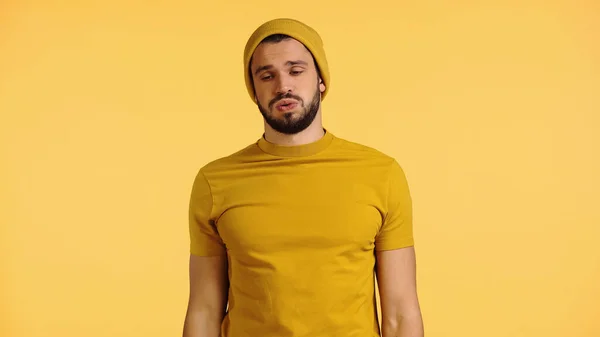 Joven cansado en gorro sombrero y camiseta hinchando mejillas aisladas en amarillo — Stock Photo