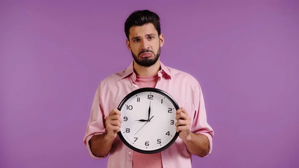 Joven molesto sosteniendo reloj aislado en púrpura - foto de stock