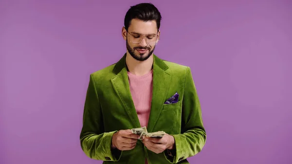 Pleased man in green jacket counting money isolated on purple — Stock Photo