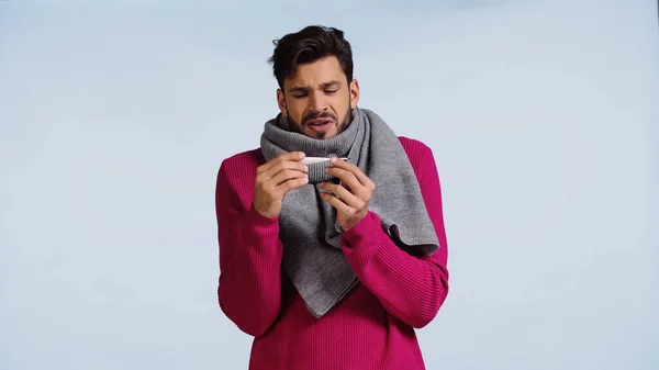 Uomo malato in maglione rosa e sciarpa guardando il termometro digitale isolato su blu — Foto stock