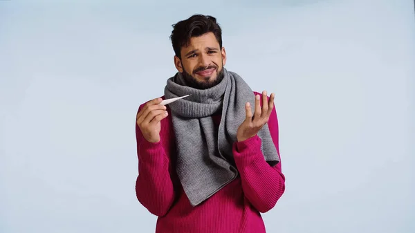 Хворий чоловік в рожевому светрі і шарфі тримає цифровий термометр ізольовано на синьому — стокове фото
