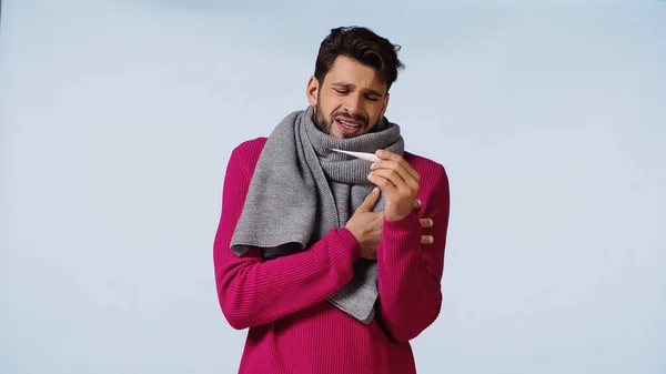 Sick man in sweater and scarf looking at digital thermometer isolated on blue — Stock Photo
