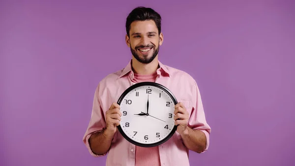 Happy young man in pink shirt holding clock isolated on purple — Stockfoto