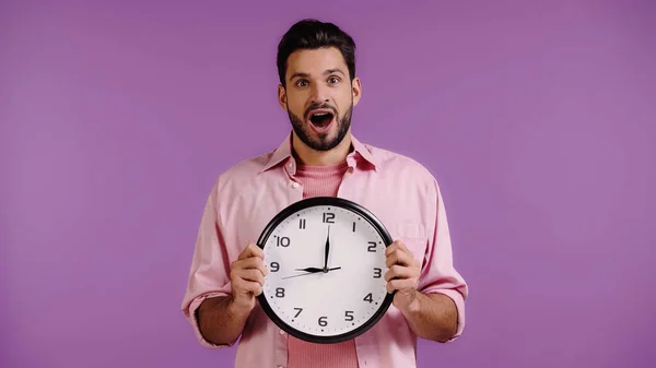 Joven excitado en camisa rosa sosteniendo reloj aislado en púrpura - foto de stock