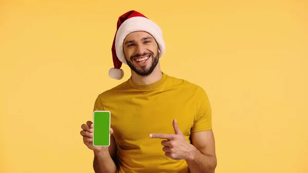 Happy man in santa hat pointing with finger at smartphone isolated on yellow — стоковое фото