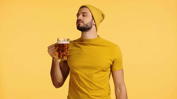 Young man in beanie hat and t-shirt holding glass mug and smelling beer isolated on yellow — Foto stock