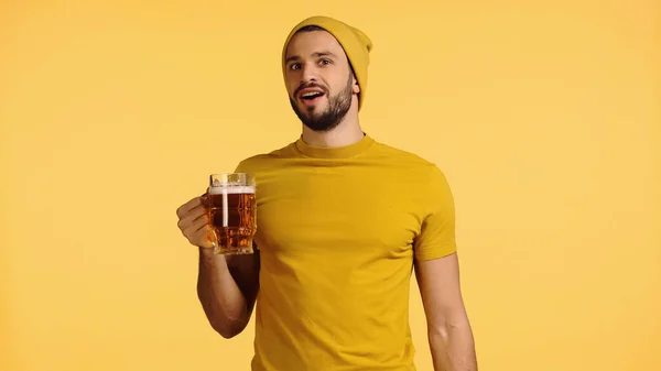 Amazed man in beanie hat and t-shirt holding glass mug with beer isolated on yellow — Stockfoto