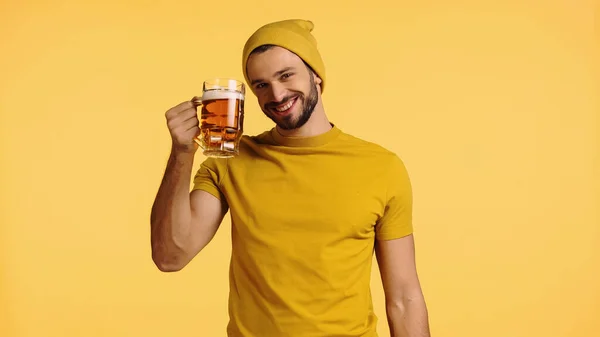 Alegre hombre en gorro sombrero y camiseta sosteniendo taza de vidrio con cerveza aislada en amarillo - foto de stock