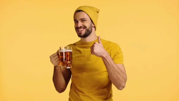 Young man in beanie hat and t-shirt holding glass mug with beer and showing thumb up isolated on yellow — Stock Photo