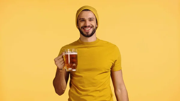 Homem feliz em chapéu gorro e t-shirt segurando caneca de vidro com cerveja isolada no amarelo — Fotografia de Stock