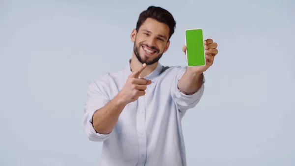 Счастливый мужчина указывает на мобильный телефон с зеленым экраном, изолированным на синем — стоковое фото