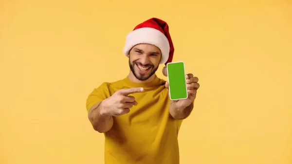 Homem feliz em santa chapéu apontando para smartphone com tela verde isolada no amarelo — Fotografia de Stock