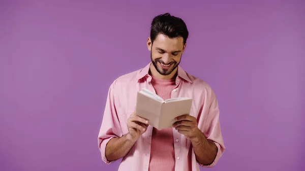 Cheerful bearded man reading book isolated on purple — Stock Photo