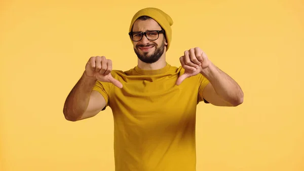 Displeased man in beanie hat and glasses showing thumbs down isolated on yellow — Stock Photo