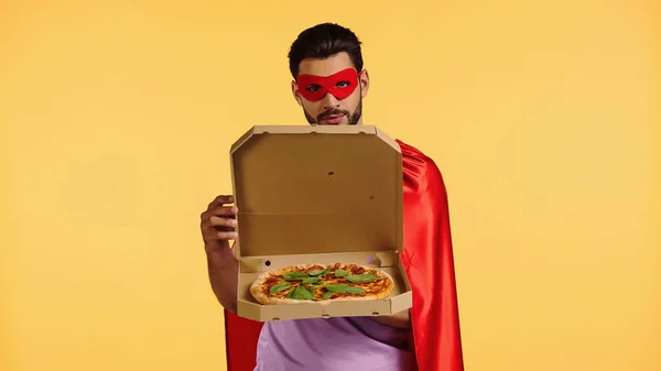 Bearded man in superhero costume opening box with pizza isolated on yellow — Stock Photo