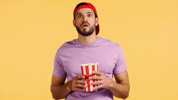 Shocked man watching movie and holding popcorn bucket isolated on yellow — стоковое фото