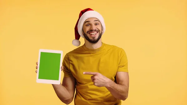 Hombre alegre en sombrero de santa señalando con el dedo a la tableta digital aislado en amarillo - foto de stock