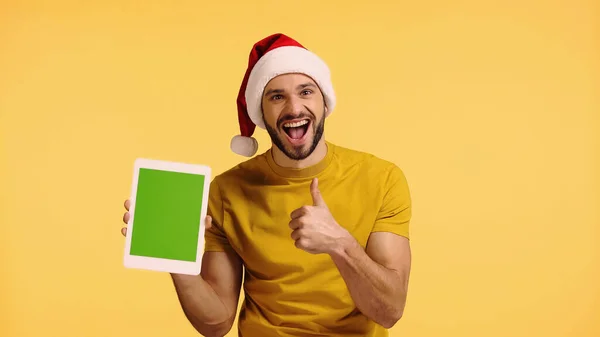 Hombre feliz en sombrero de santa celebración tableta digital y mostrando el pulgar hacia arriba aislado en amarillo - foto de stock