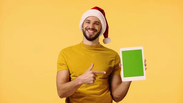 Hombre feliz en sombrero de santa señalando con el dedo a la tableta digital aislado en amarillo - foto de stock