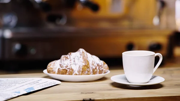 Taza de café cerca de croissant en el plato y periódico en la mesa - foto de stock