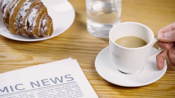 Cropped view of man taking coffee with milk near newspaper and croissant — Stock Photo