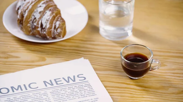 Tasse en verre d'espresso près du journal, de l'eau et du croissant — Photo de stock