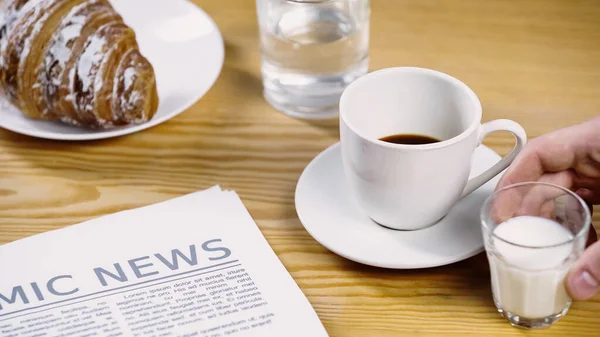 Cropped view of man taking glass of milk near cup of coffee, newspaper and croissant — Stock Photo