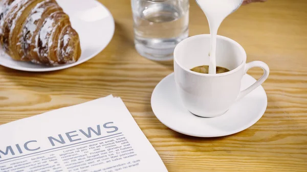 Milch gießt auf Tasse Kaffee in der Nähe von Zeitung und leckerem Croissant — Stockfoto