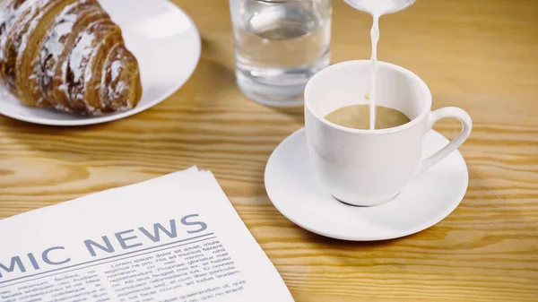 Milk pouring onto cup of coffee near newspaper and croissant — Stock Photo