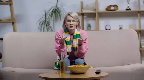 Bouleversé femme en écharpe rayée pointant avec les mains tout en regardant le jeu de sport à la télévision à la maison — Photo de stock