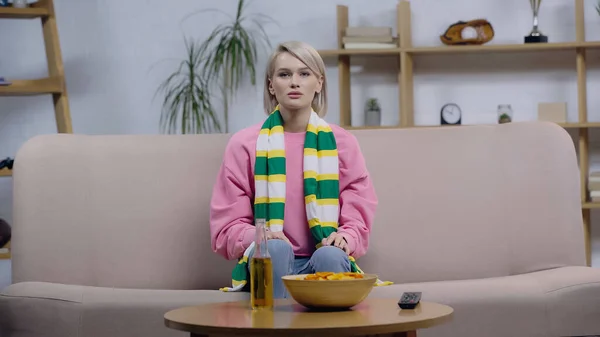 Frustrated sport fan woman in striped scarf watching match on tv near chips and bottle of beer — Fotografia de Stock