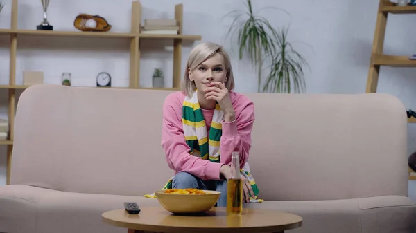 Displeased woman in striped scarf watching sport championship on sofa near chips and beer — Stockfoto