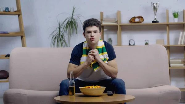 Worried sport fan in striped scarf watching championship on sofa near beer and chips — Fotografia de Stock