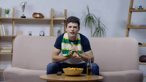 Excited sport fan sitting on sofa near chips and beer and watching game on tv — Foto stock