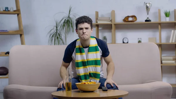 Joven tenso en bufanda rayada viendo deporte juego en el sofá cerca de la cerveza y patatas fritas - foto de stock