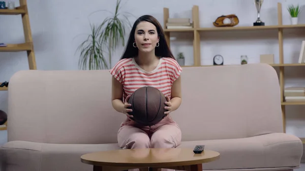 Positivo e concentrado fã de esporte mulher assistindo jogo de basquete na tv enquanto sentado no sofá com bola — Fotografia de Stock
