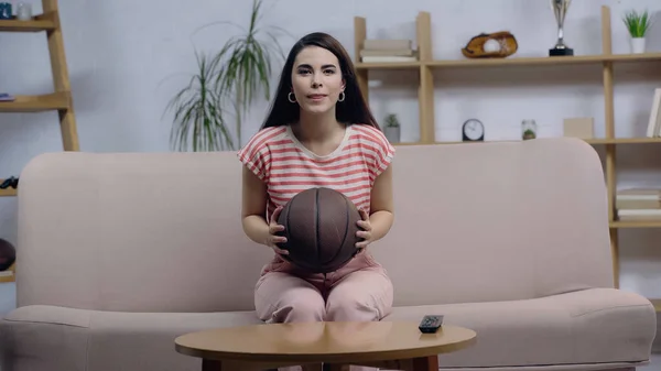 Positive and concentrated sport fan woman watching basketball match on tv while sitting on couch with ball — Stockfoto