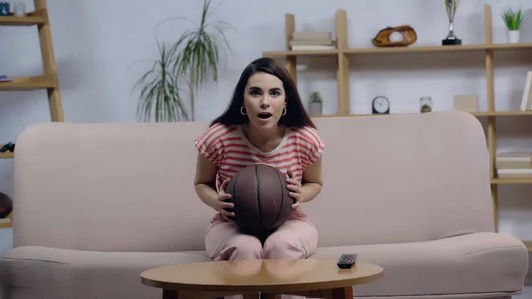 Stupito sport fan donna seduta sul divano con la bocca aperta mentre si guarda partita di basket — Foto stock