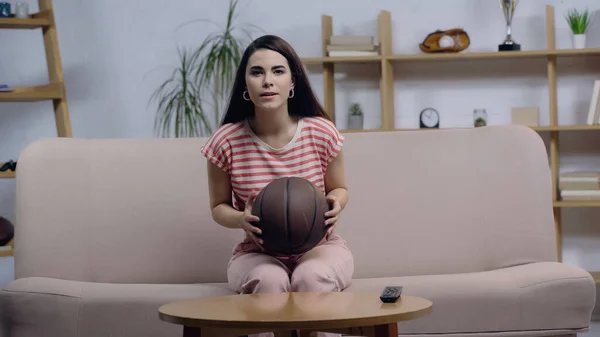 Ventilador de baloncesto enfocado viendo el juego en casa mientras está sentado en el sofá con la pelota - foto de stock