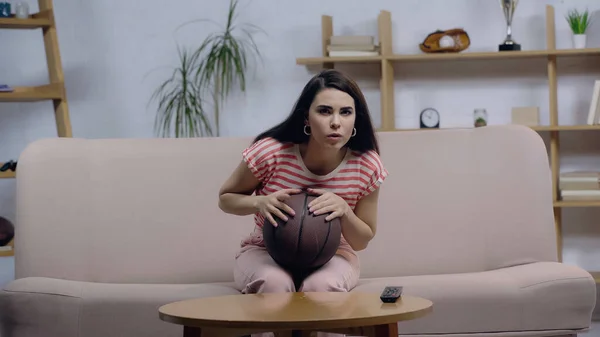 Concentrated woman sitting on couch with ball while watching basketball game on tv — Stock Photo