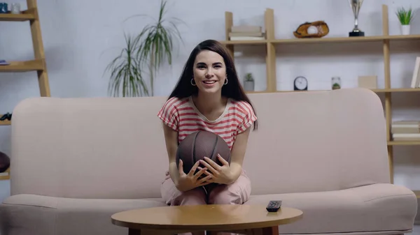 Mujer alegre sentada en el sofá con la pelota y viendo el partido de baloncesto en la televisión - foto de stock