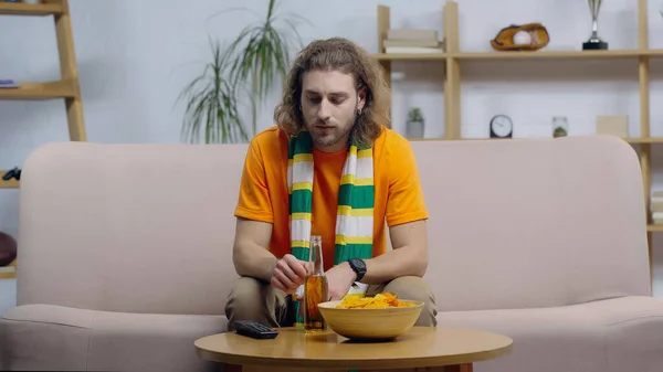 Sad sport fan in striped scarf sitting on couch near tv remote controller, beer and chips on coffee table — Stockfoto