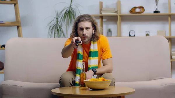 Fan de sport en écharpe rayée en cliquant sur les chaînes de télévision tout en étant assis près de la bière et des chips sur la table basse — Photo de stock