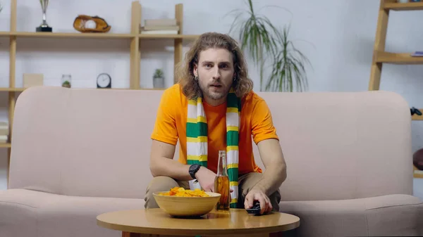 Young sport fan clicking tv channels while sitting on couch at home near chips and beer — Fotografia de Stock