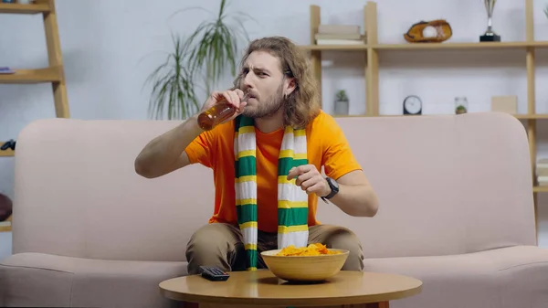 Young man in striped scarf drinking beer while watching sport match on tv at home — Stock Photo