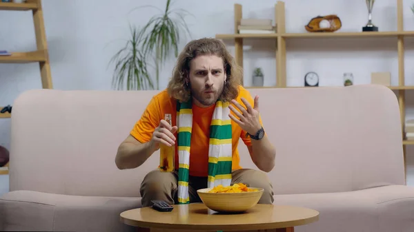 Worried sport fan in striped scarf holding bottle of beer and gesturing while watching match on tv — Foto stock