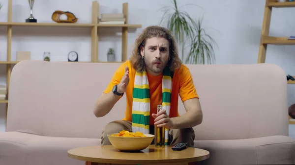 Worried man in striped scarf pointing with hand while watching sport competition near beer and chips — Stock Photo