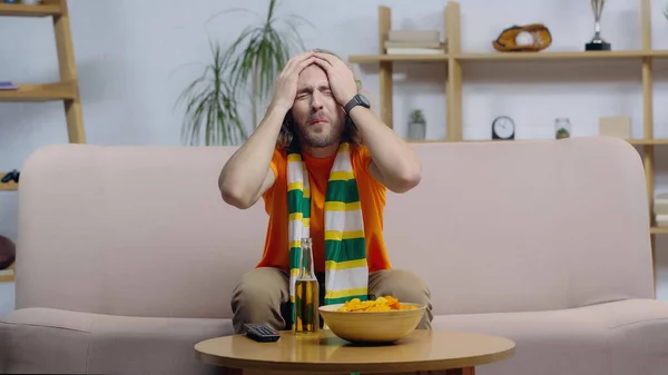 Ventilateur de sport bouleversé dans l'écharpe rayée tenant la main sur la tête près de la télécommande de télévision, bière et chips sur la table basse — Photo de stock