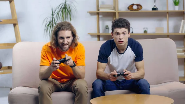 KYIV, UKRAINE - DECEMBER 22, 2021: excited man playing video game with concentrated friend on couch at home — Stock Photo